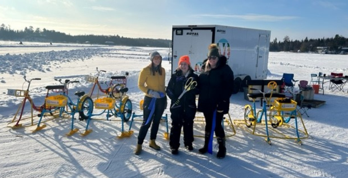 Three people standing together in snowy area with utility trailer and ice bikes in the background